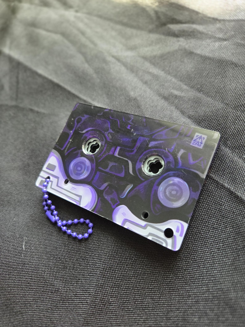 May include: A rectangular acrylic keychain shaped like a cassette tape featuring a dark purple and black abstract pattern. The design includes two circular cutouts, a small logo in the corner, and a purple ball chain attached to the side.