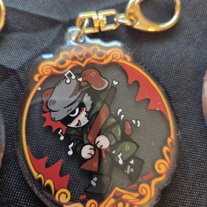 Op de afbeelding: Een heldere acryl sleutelhanger met een stripfiguur in een militaire outfit, die een geweer vasthoudt. Het ontwerp is geplaatst in een rode en gouden decoratieve rand, met een goudkleurige sluiting.