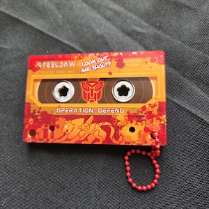 May include: A rectangular orange and red cassette tape keychain featuring the text STEELJAW, LOOK OUT AND SHOUT!, and OPERATION: DEFEND. The design includes a red Autobot symbol, a stylized lion graphic, and a red ball chain attached to the corner.