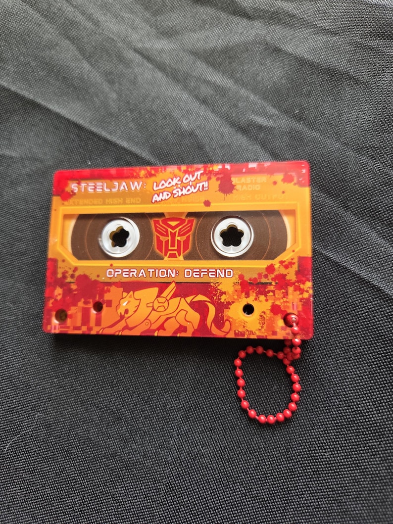 May include: A rectangular orange and red cassette tape keychain featuring the text STEELJAW, LOOK OUT AND SHOUT!, and OPERATION: DEFEND. The design includes a red Autobot symbol, a stylized lion graphic, and a red ball chain attached to the corner.