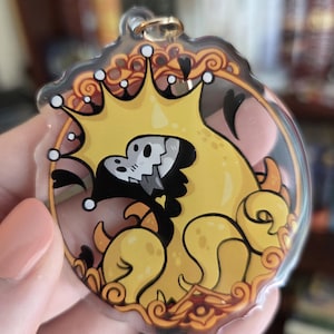 Op de afbeelding: Een heldere acryl sleutelhanger met een gele cartoonfiguur met een kroon en een schedelachtig gezicht. Het ontwerp is omlijnd in zwart en oranje, met een goudkleurige metalen ring voor bevestiging. De sleutelhanger wordt in een hand gehouden.
