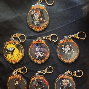 May include: Collection of seven acrylic keychains with gold-colored hardware. Each keychain features a unique cartoon character design within an oval frame with ornate gold detailing. The backgrounds vary, with some featuring red or yellow accents.