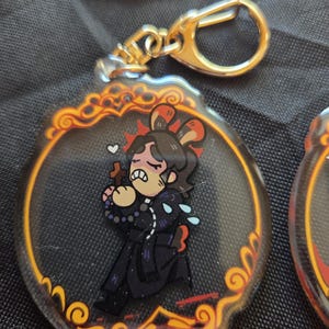 Op de afbeelding: Een heldere acryl sleutelhanger met een goudkleurige sluiting. De sleutelhanger heeft een cartoon illustratie van een persoon in een zwarte outfit met een kruis, een rode hoofdband en een bedroefde uitdrukking. De rand is een decoratief oranje en goudkleurig ontwerp.