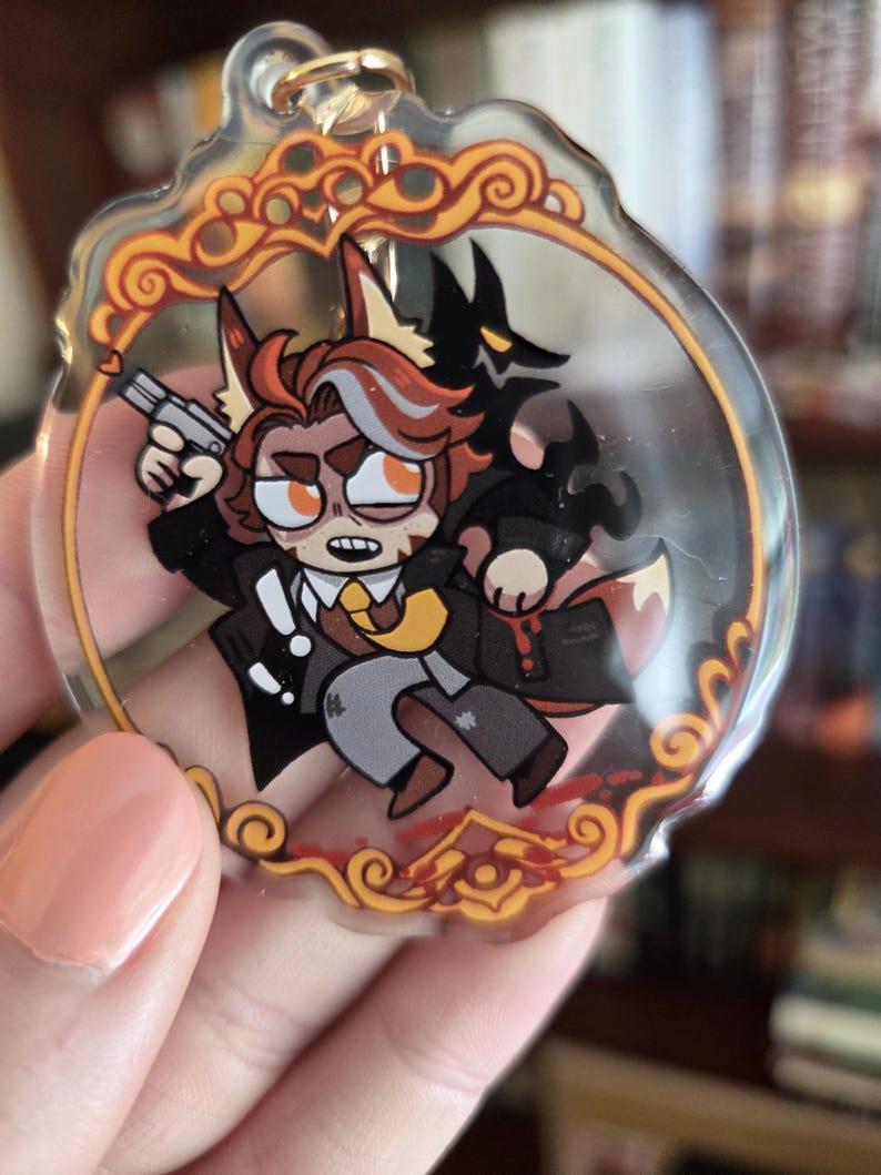 Op de afbeelding: Een heldere acryl sleutelhanger met een cartoon vos karakter in een pak met een pistool, met een zwart schaduwfiguur achter hem. De sleutelhanger heeft een gouden rand met een sierlijk ontwerp.
