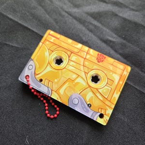 May include: A rectangular yellow cassette tape keychain featuring a red Autobot symbol. The design includes metallic gray accents on the corners and two circular cutouts in the center. A small red ball chain is attached to the side of the object.