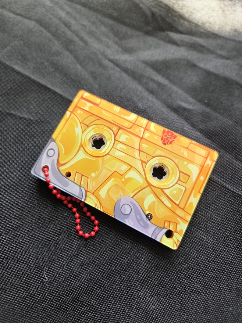 May include: A rectangular yellow cassette tape keychain featuring a red Autobot symbol. The design includes metallic gray accents on the corners and two circular cutouts in the center. A small red ball chain is attached to the side of the object.