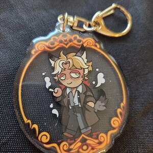 Op de afbeelding: Een heldere acryl sleutelhanger met een stripfiguur met kattenoren, gekleed in een bruine jas en met een sigaret. De sleutelhanger heeft een goudkleurige sluiting en een decoratieve rand met oranje en gouden accenten.