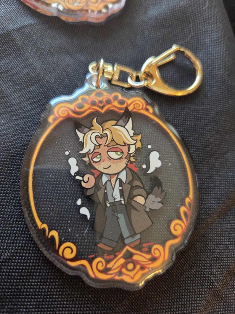 Op de afbeelding: Een heldere acryl sleutelhanger met een stripfiguur met kattenoren, gekleed in een bruine jas en met een sigaret. De sleutelhanger heeft een goudkleurige sluiting en een decoratieve rand met oranje en gouden accenten.