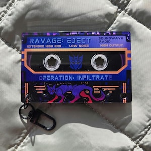 May include: A rectangular acrylic keychain shaped like a cassette tape with a purple and black color scheme. It features the text RAVAGE: EJECT, SOUNDWAVE RADIO, and OPERATION: INFILTRATE, along with a purple panther graphic and a metal lobster clasp.