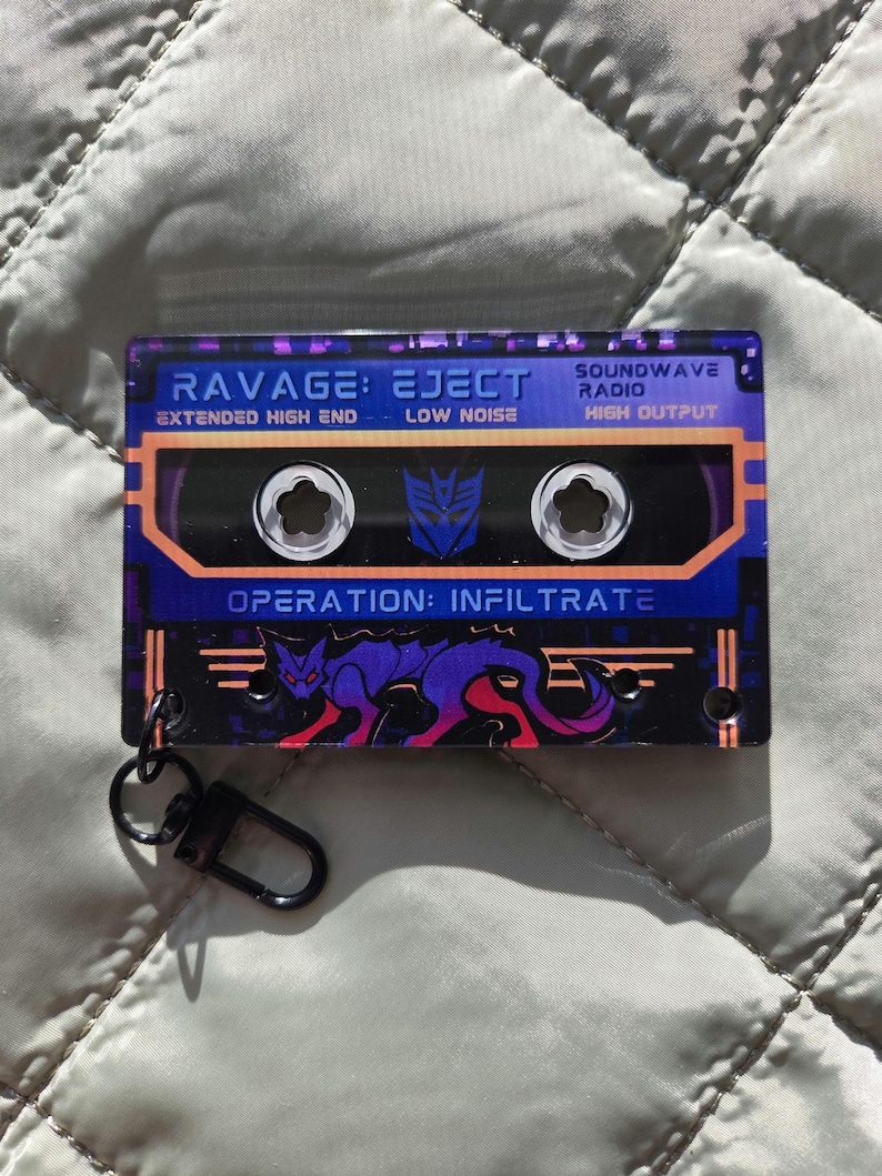 May include: A rectangular acrylic keychain shaped like a cassette tape with a purple and black color scheme. It features the text RAVAGE: EJECT, SOUNDWAVE RADIO, and OPERATION: INFILTRATE, along with a purple panther graphic and a metal lobster clasp.