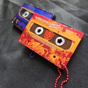 May include: Two cassette tape-shaped accessories with attached ball chains. The front one is orange and red with the text "STEELJAW: LOOK OUT AND SHOUT!!" and "OPERATION: DEFEND". The back one is purple and blue with the text "EJECT" and "OPERATION".