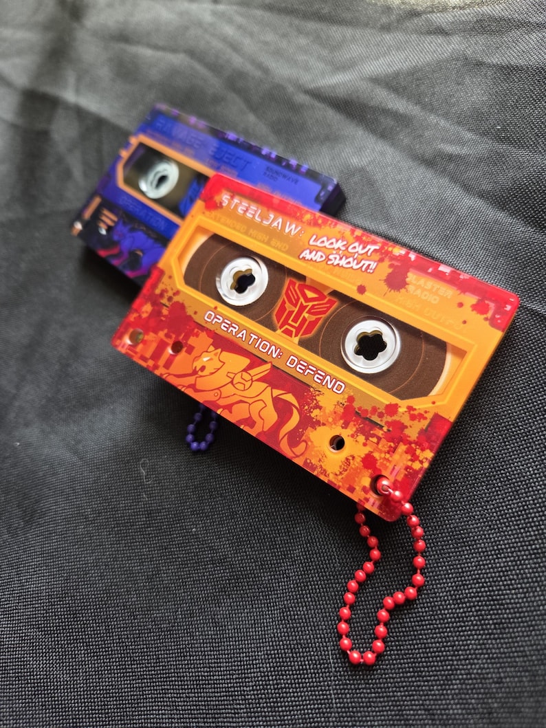 May include: Two cassette tape-shaped accessories with attached ball chains. The front one is orange and red with the text "STEELJAW: LOOK OUT AND SHOUT!!" and "OPERATION: DEFEND". The back one is purple and blue with the text "EJECT" and "OPERATION".