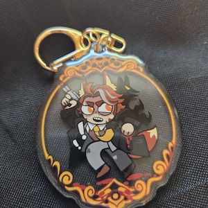 Op de afbeelding: Een heldere acryl sleutelhanger met een stripfiguur met vosachtige kenmerken, die een pistool vasthoudt. Het personage draagt een pak met een gele stropdas en een zwart silhouet van een wolf staat achter hem. De sleutelhanger heeft een goudkleurige sluiting.