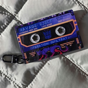 May include: A rectangular acrylic keychain shaped like a cassette tape with a purple and orange color scheme. It features the text RAVAGE: EJECT, SOUNDWAVE RADIO, and OPERATION: INFILTRATE, along with a graphic of a purple panther and a Decepticon logo.