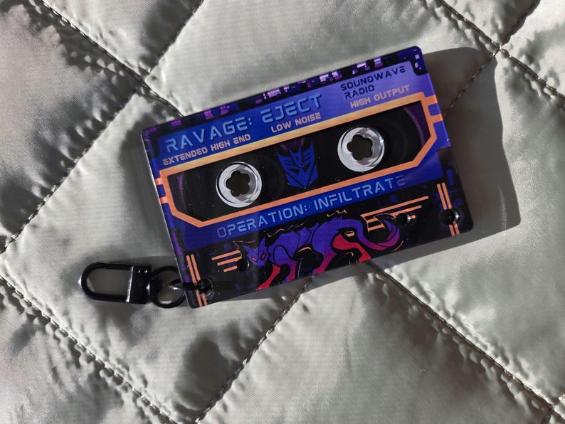 May include: A rectangular acrylic keychain shaped like a cassette tape with a purple and orange color scheme. It features the text RAVAGE: EJECT, SOUNDWAVE RADIO, and OPERATION: INFILTRATE, along with a graphic of a purple panther and a Decepticon logo.