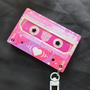 May include: A pink cassette tape keychain featuring the text ROSANNA EJECT, OPERATION SHINE, and a central Autobot symbol. The design includes white heart graphics, star patterns, and a white plastic lobster clasp attached to the bottom corner.