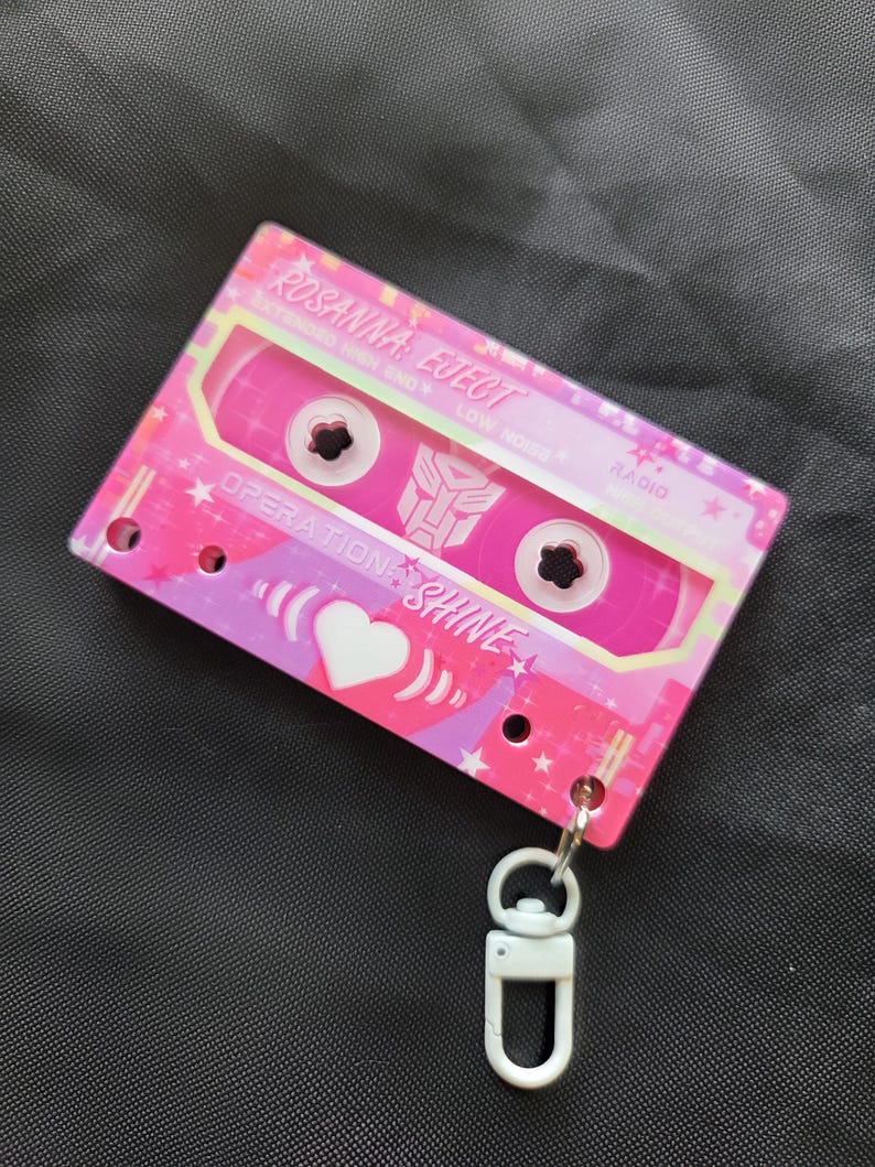 May include: A pink cassette tape keychain featuring the text ROSANNA EJECT, OPERATION SHINE, and a central Autobot symbol. The design includes white heart graphics, star patterns, and a white plastic lobster clasp attached to the bottom corner.