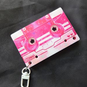 May include: A rectangular keychain shaped like a cassette tape with a pink and white color scheme, featuring a glittery finish, small heart icons, and the name ROSANNA printed in black. It includes a white plastic swivel lobster clasp attachment.