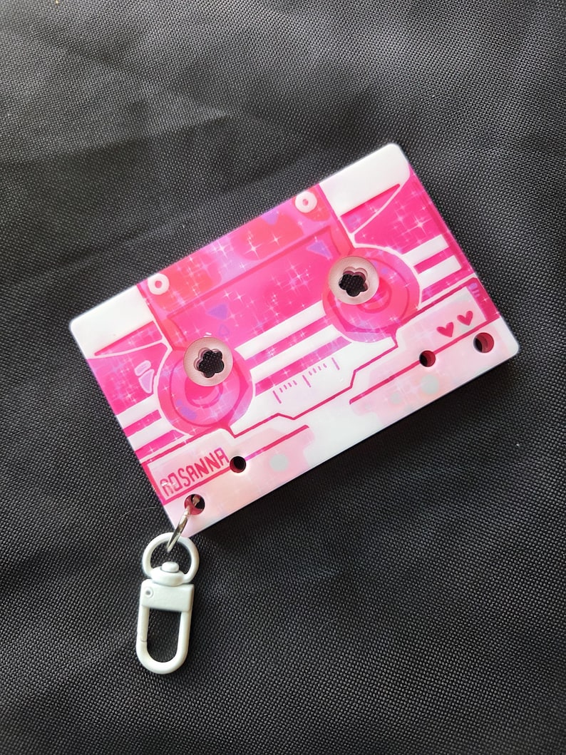 May include: A rectangular keychain shaped like a cassette tape with a pink and white color scheme, featuring a glittery finish, small heart icons, and the name ROSANNA printed in black. It includes a white plastic swivel lobster clasp attachment.