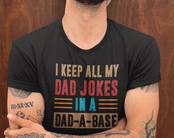 May include: Black t-shirt with the text "I KEEP ALL MY DAD JOKES IN A DAD-A-BASE" in a retro color scheme. The person in the image is wearing a light gray beanie and has tattoos on their arms.