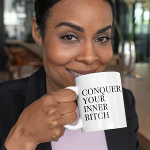 以下が含まれることがあります： 「CONQUER YOUR INNER BITCH」の黒い文字がプリントされた白いセラミックマグカップを人が持っています。マグカップはシンプルなデザインで、大胆なメッセージが書かれています。人は微笑んでいます。
