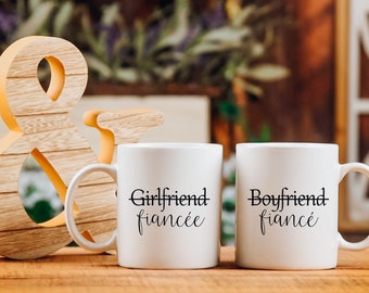 Op de afbeelding: Twee witte keramische mokken met zwarte tekst "Girlfriend fiancée" en "Boyfriend fiancé". Een houten ampersand en een rustieke achtergrond met bloemen zijn ook zichtbaar.