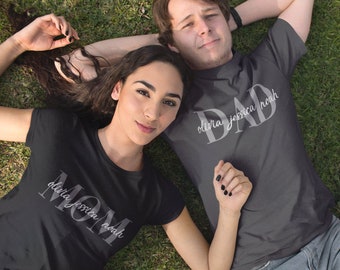 Peut inclure: Un couple portant des t-shirts noirs assortis avec les mots "MOM" et "DAD" en texte blanc. Les t-shirts comprennent également les noms "Olivia Jessica Noah" en texte blanc plus petit. Le couple est allongé sur le dos dans un champ herbeux.