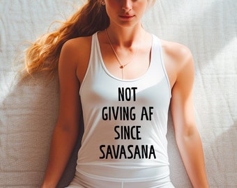 Può includere: Una persona con una canotta bianca e pantaloni bianchi è seduta in una posa yoga. La canotta ha il testo "NOT GIVING AF SINCE SAVASANA" stampato in nero. La persona ha lunghi capelli biondi ed è seduta su un tappetino da yoga grigio chiaro.