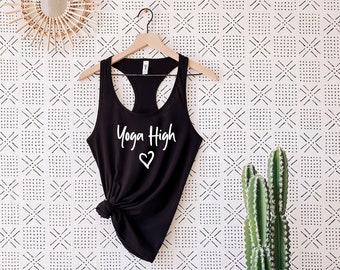 May include: Black tank top with the text "Yoga High" and a heart symbol in white lettering. The tank top is hanging on a wooden hanger against a white wall with a black geometric pattern.