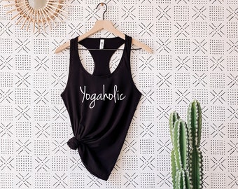 May include: Black racerback vest with the word "Yogaholic" in white script. The vest is hanging on a wooden hanger against a patterned white and black background. A cactus in a white pot sits on a wooden stump.