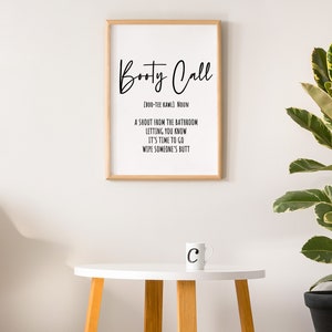 May include: A framed print with the words "Booty Call" in a stylish font, with a definition below. A white mug with the letter "C" sits on a small white table with wooden legs. A potted plant is in the background.