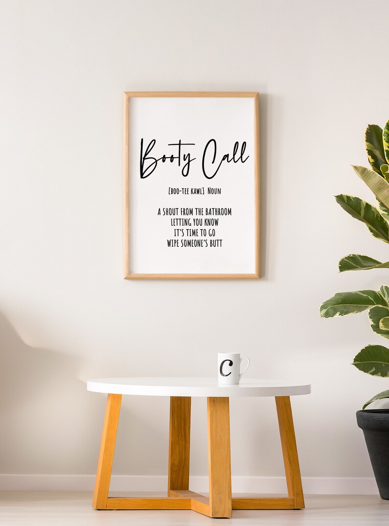 May include: A framed print with the words "Booty Call" in a stylish font, with a definition below. A white mug with the letter "C" sits on a small white table with wooden legs. A potted plant is in the background.