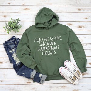 K&ouml;nnte beinhalten: Ein olivgr&uuml;ner Kapuzenpullover mit dem wei&szlig;en Text "I RUN ON CAFFEINE, SARCASM & INAPPROPRIATE THOUGHTS". Dazu abgebildet sind zerrissene blaue Jeans und wei&szlig;e Turnschuhe, alles auf einer hellen Holzmaserung.