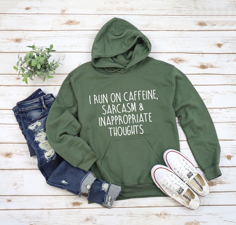 K&ouml;nnte beinhalten: Ein olivgr&uuml;ner Kapuzenpullover mit dem wei&szlig;en Text "I RUN ON CAFFEINE, SARCASM & INAPPROPRIATE THOUGHTS". Dazu abgebildet sind zerrissene blaue Jeans und wei&szlig;e Turnschuhe, alles auf einer hellen Holzmaserung.
