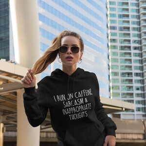 K&ouml;nnte beinhalten: Schwarzer Kapuzenpullover mit dem wei&szlig;en Text "I RUN ON CAFFEINE, SARCASM & INAPPROPRIATE THOUGHTS". Der Hoodie hat eine Vordertasche und eine Kapuze mit Kordelzug. Die Person im Bild tr&auml;gt eine Sonnenbrille und hellgr&uuml;ne Leggings.