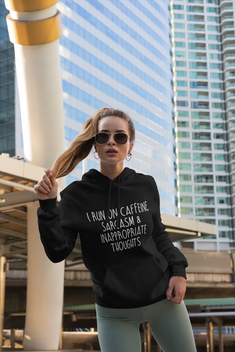 K&ouml;nnte beinhalten: Schwarzer Kapuzenpullover mit dem wei&szlig;en Text "I RUN ON CAFFEINE, SARCASM & INAPPROPRIATE THOUGHTS". Der Hoodie hat eine Vordertasche und eine Kapuze mit Kordelzug. Die Person im Bild tr&auml;gt eine Sonnenbrille und hellgr&uuml;ne Leggings.