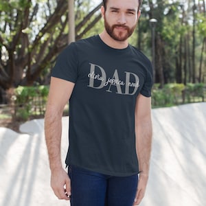 May include: A dark blue t-shirt with the word "DAD" in large white letters. The names "Olivia, Jessica, Noah" are written in smaller white letters below the word "DAD".