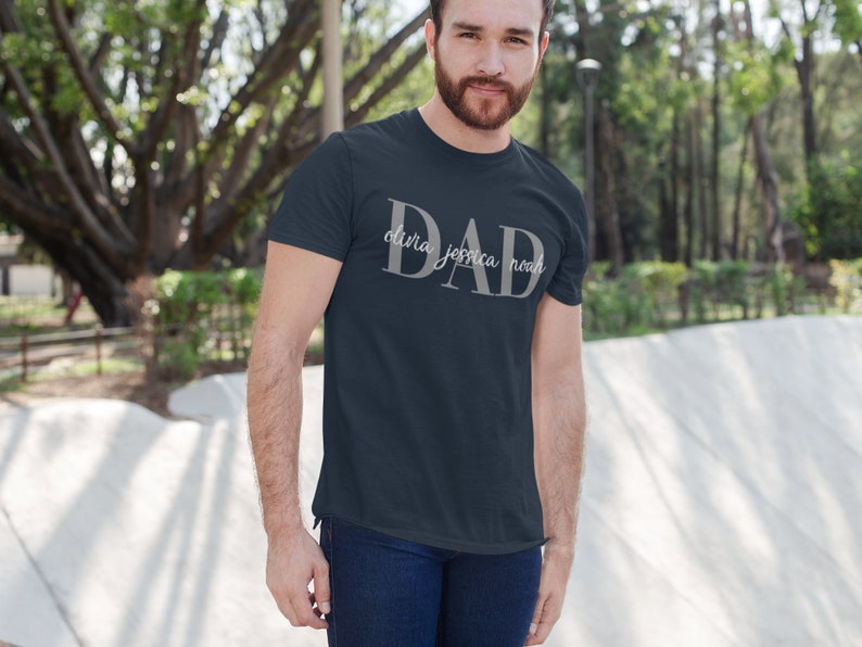 May include: A dark blue t-shirt with the word "DAD" in large white letters. The names "Olivia, Jessica, Noah" are written in smaller white letters below the word "DAD".