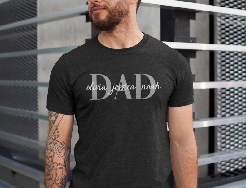 May include: A dark heather grey t-shirt with the word "DAD" in large white letters. Below the word "DAD" are the names "Olivia Jessica Noah" in smaller white letters.