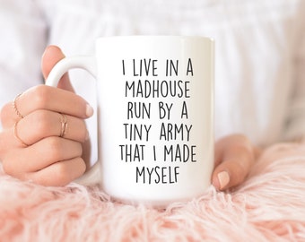 May include: White ceramic mug with the text "I LIVE IN A MADHOUSE RUN BY A TINY ARMY THAT I MADE MYSELF" in black. The mug is held by a person with multiple gold rings on their fingers.