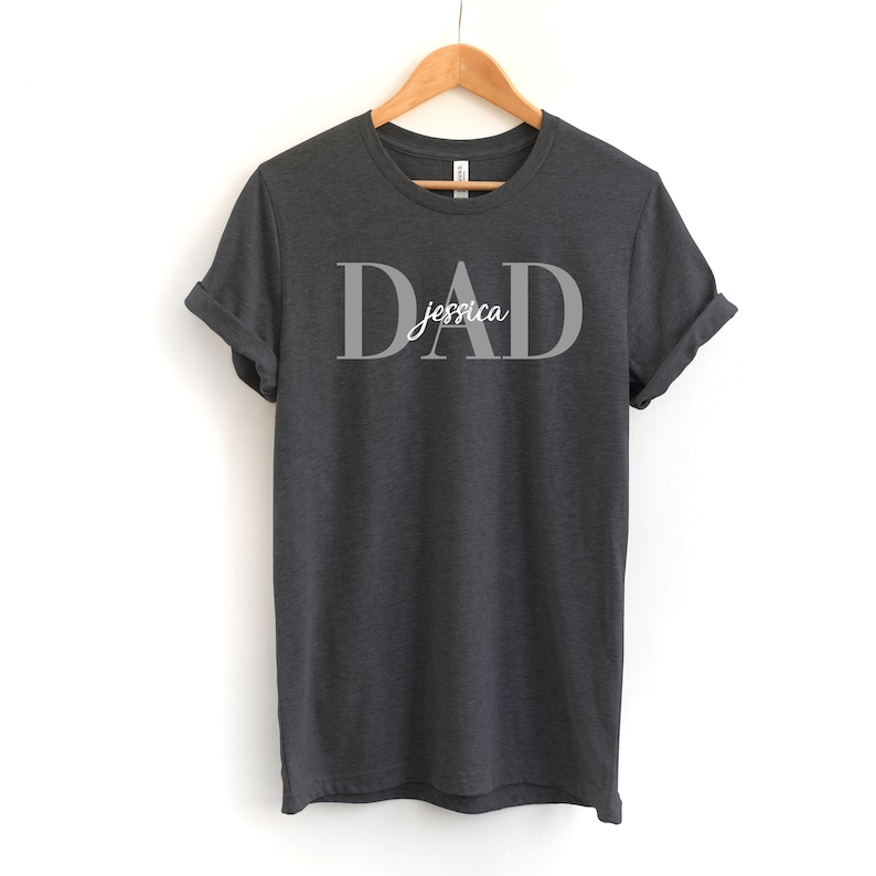 May include: Dark gray t-shirt with the word "DAD" in large, gray letters and the name "Jessica" written in a smaller, white font underneath.