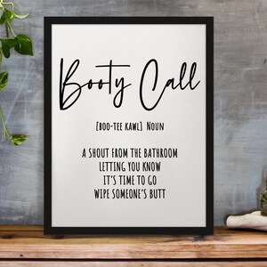 May include: Black framed art print with the words "Booty Call" in a cursive font. The definition of "Booty Call" is listed below the title. The print is displayed on a wooden surface.