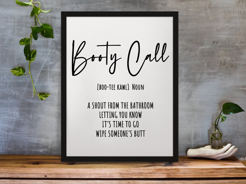 May include: Black framed art print with the words "Booty Call" in a cursive font. The definition of "Booty Call" is listed below the title. The print is displayed on a wooden surface.