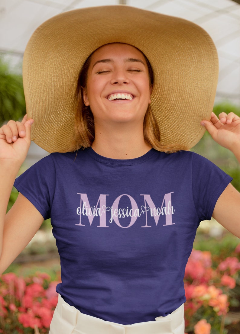 May include: Navy blue t-shirt with the word "MOM" in light purple, with the names Olivia, Jessica, and Noah below. The shirt is worn by a person wearing a large straw hat.
