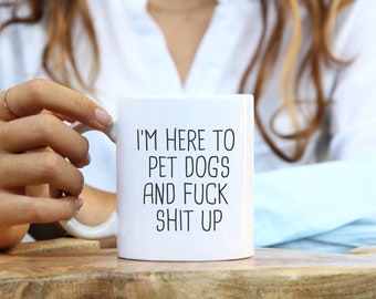 May include: White ceramic mug with the text "I'M HERE TO PET DOGS AND FUCK SHIT UP" in black, sans-serif font. The mug has a white handle and is held by a person. The mug is on a wooden surface.