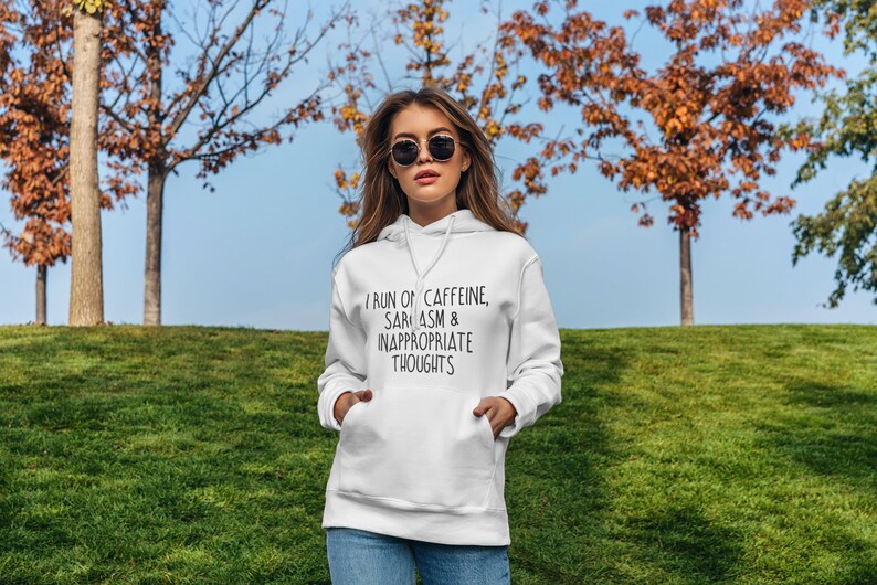 K&ouml;nnte beinhalten: Ein wei&szlig;er Kapuzenpullover mit dem Text "I RUN ON CAFFEINE, SARASM & INAPPROPRIATE THOUGHTS" in Schwarz. Der Hoodie hat eine Vordertasche und eine Kapuze mit Kordelzug. Die Person tr&auml;gt eine Sonnenbrille und steht auf einem grasbewachsenen H&uuml;gel.