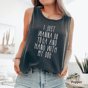 May include: A dark gray tank top with the text "I JUST WANNA DO YOGA AND HANG WITH MY DOG" in white. The tank top is sleeveless and has a rounded neckline. The model is wearing blue jeans.