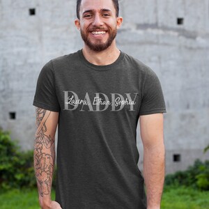 May include: A dark gray t-shirt with the word "DADDY" in white lettering. The names "Laura, Ethan, Sophia" are written in smaller white lettering below the word "DADDY".