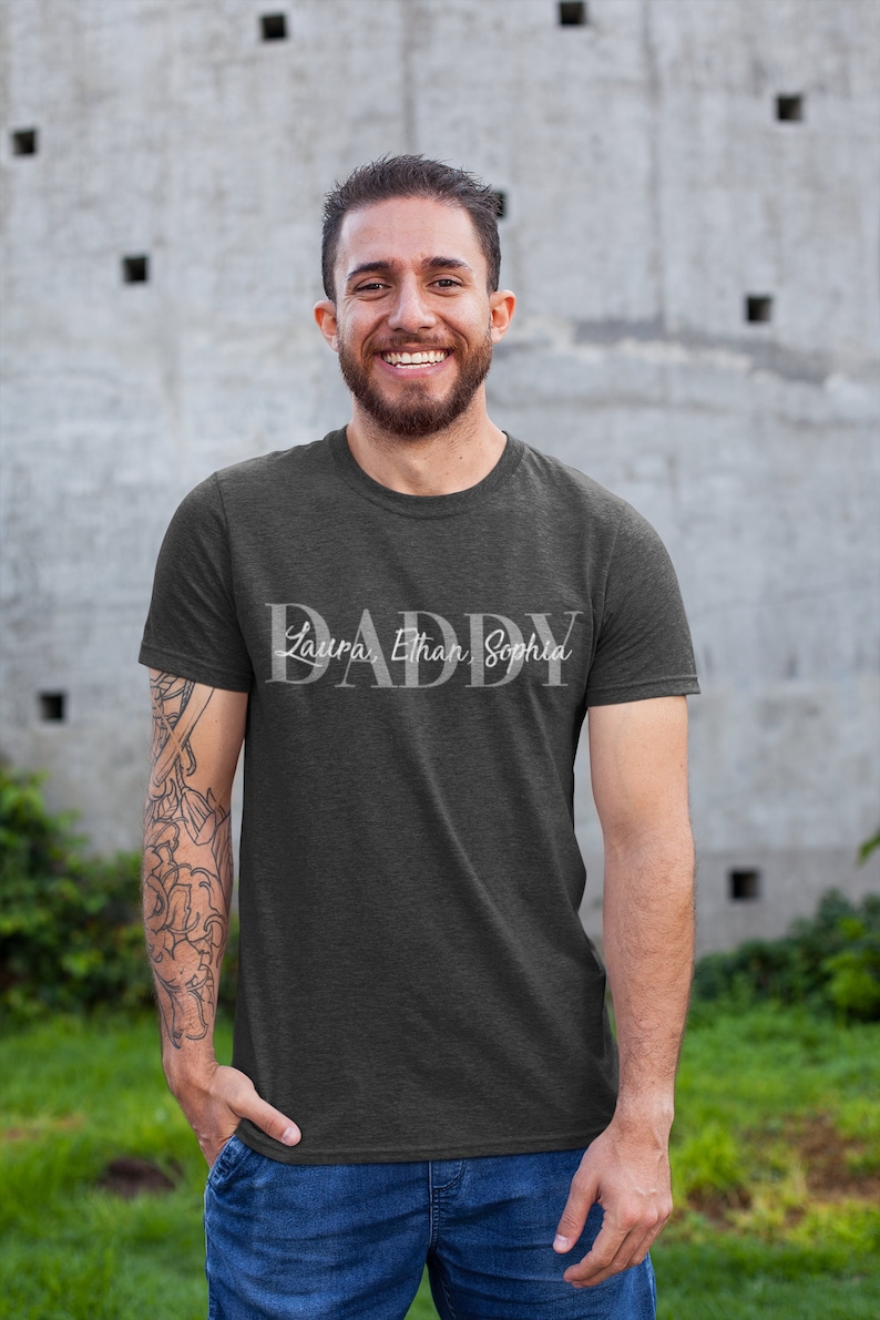 May include: A dark gray t-shirt with the word "DADDY" in white lettering. The names "Laura, Ethan, Sophia" are written in smaller white lettering below the word "DADDY".