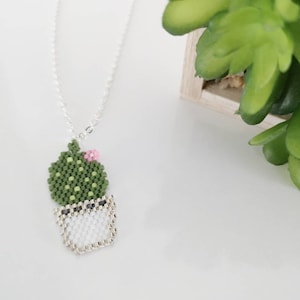 May include: A silver chain necklace with a beaded cactus charm. The cactus is green with a pink flower and is potted in white.